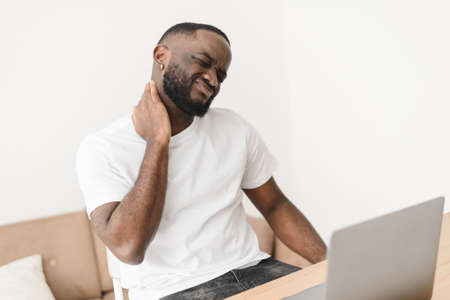 Portrait of a tense handsome black man with neck pain. Man working on business project using laptop suffering from neck painの写真素材