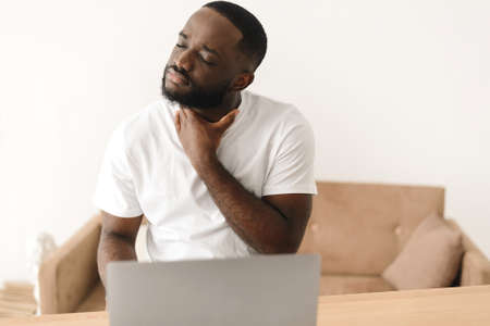 A black man works remotely with a laptop from home and suffers from a sore throat. An African American man begins to get sick and feel a sore throat.の写真素材