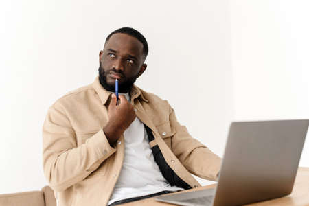 A black young man sitting at a table studying distance learning improves knowledge, looks thoughtfully to the side, holding a pen to his mouth. E-learning conceptの写真素材