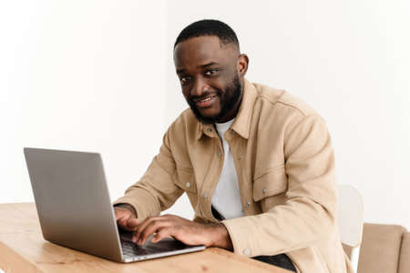 Portrait of smiling african american man looking at camera while sitting at table in home office and working on laptop. Happy black freelancer posing at workplace at home.の写真素材