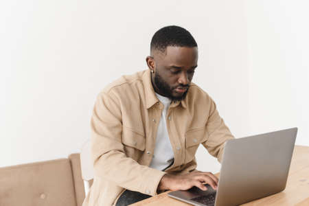 Concentrated focused young african freelancer sits at the table and looks into the laptop. Serious black businessman working online with computer data watching a webinar thinking about solving a problem.の写真素材