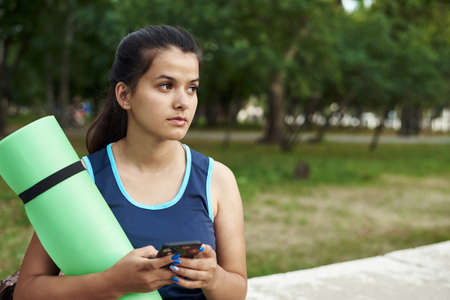 Close-up of young sporty woman in blue sports clothing holding exercise mat and looking at her mobile phone in her hand while standing outdoorsの写真素材