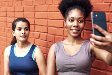 Image of amazing young athletic women taking a selfie against a brick wall. Smiling black women spend time together after sports workoutの写真素材