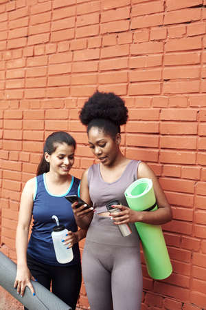 Image of amazing young athletic women in front of a brick wall, looking into the phone. Two female friends laugh at the social media postの写真素材