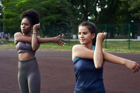 Two fit smiling young woman working out together in a park doing stretching exercise and hand extensions to improve supplenessの写真素材