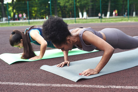 Two athletic female friends do push-ups on the exercise mat at the stadium in the park. Muscle load.