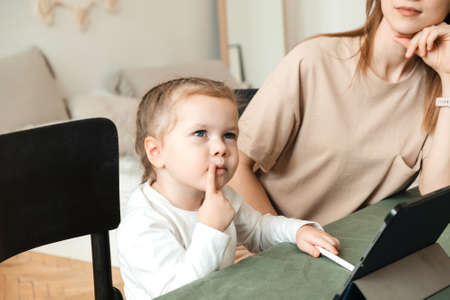 A little girl is thinking how to do tasks on a tablet at home or complete a game. Mom helps to do homework for her daughters kindergarten.の写真素材