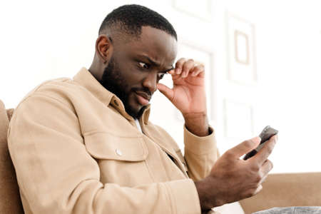Displeased african american man using mobile phone has problems with mobile app while sitting on sofa at home. Smartphone problem. Displeased phone user reading negative news onlineの写真素材