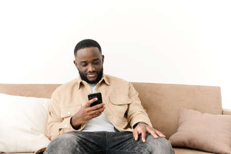 Happy bearded afro american freelancer working using phone while sitting on sofa at his home. Concept of young people working on mobile devicesの写真素材