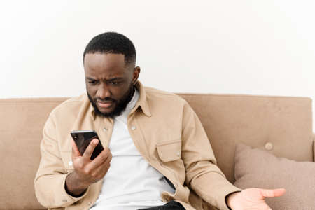 Annoyed african american man yelling at phone while sitting on sofa at home. Angry man looks at the phone while reading bad news. Business problems.の写真素材