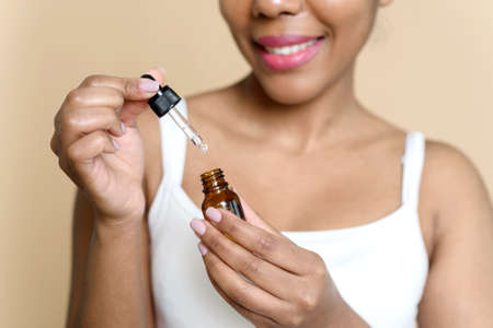 Beautiful woman applying hyaluronic acid for facial skin care and treatment with collagen moisturizer. Close up african american beauty holding glass bottle with moisturizing serum on beige backgroundの写真素材