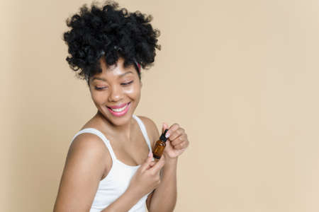 Portrait of a beautiful black woman holding a bottle of moisturizing serum on a beige background. African American young woman smiling while holding a jar of oil.の写真素材