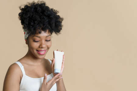 Bottle of cosmetic cream with a mockup in the hands of an african american female model on a beige background. Lovely black woman advertising moisturizerの写真素材