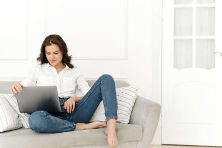 Young positive woman using laptop, checking news on internet, sitting on sofa, working remotelyの写真素材