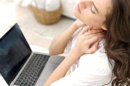 Tired young woman with neck pain working in laptop at home office. Tired young woman touching her stiff neck, feeling joint pain, backache, massaging tense musclesの写真素材