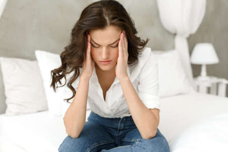 Unhappy woman touching her head, sitting on the bed at home, having a headache. Young frustrated woman thinks about problems, feels lonely and sadの写真素材
