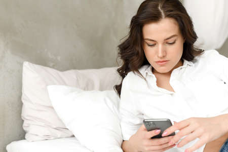 Happy young woman with mobile phone lying on the sofa. Young happy woman lies down on bed and browses social networks at home on smartphone.の写真素材