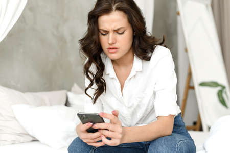 A young woman scrolls through her social media feed and sees some bad news. Caucasian female model sits on the bed and at home and worries, reading about problems, spam in the mailの写真素材