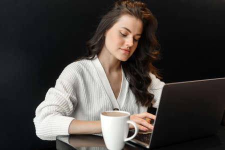 Smiling woman is sitting in the dining room working on a laptop. Nearby is a glass of coffee or cocoa. Business woman checks email on laptop.の写真素材