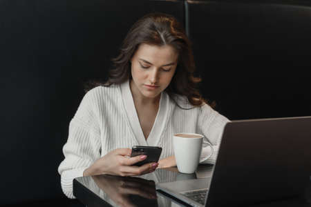 A young concentrated woman in a cafe reads a text message or news from her mobile phone, sits at a table in a cafe with a laptop.の写真素材