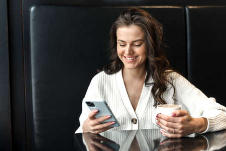 Young beautiful smiling woman holding coffee cup and looking at smartphone while sitting in dining room. A happy university student using a mobile phone reads the news. Business woman drinking coffee and smiling in a dark interior of a coffee shopの写真素材
