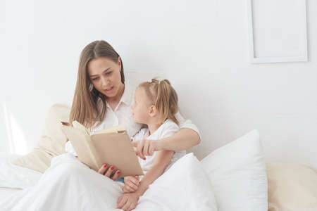 Close-up mom and little daughter are resting in a cozy bed, reading a funny interesting childrens book togetherの写真素材