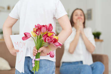 Cute little girl hides flowers and a gift card for her mother behind her back, close-up. Mom is looking forward to the gift.の写真素材