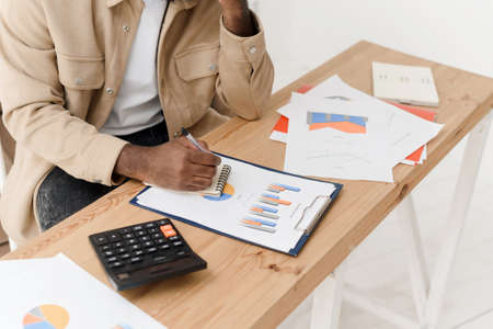 Close-up of an African American man calculating with a calculator, managing household finances, or keeping company financial records at home. Black man making calculations on a calculator, paying bills, taking into account taxes or expenses.の写真素材