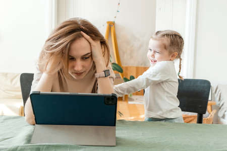 Mother trying to work from home on a tablet with her daughter. Quarantine and remote work during the outbreak. The child makes noise and interferes with the woman at work from home.の写真素材