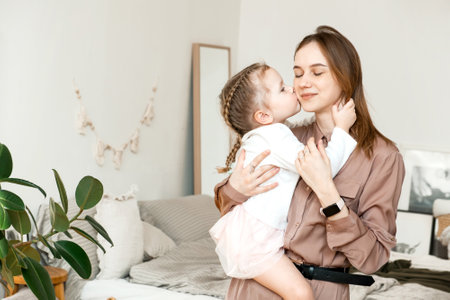 A little girl kisses her mother on the cheek at home in a modern bright bedroom. The concept of parenthood, love and careの写真素材