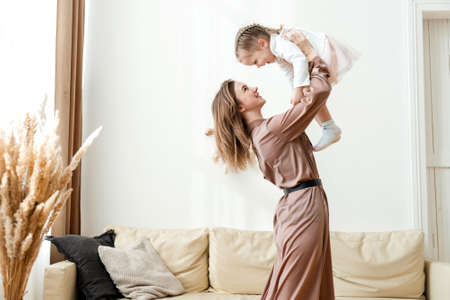 Smiling mother lifts happy little daughter into the air in the living room and plays with her.の写真素材