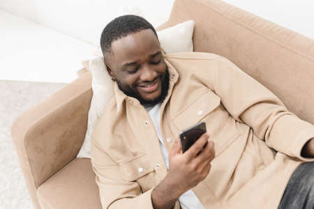Black guy using smartphone, browsing internet and texting while lying on sofa at home. Network social media application. Mobile connection.の写真素材