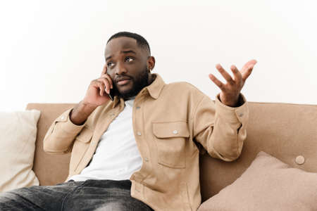 Close-up of an annoyed African American young man sitting on a couch talking on a cell phone. Angry black young man talking on smartphone, arguing with girlfriend or serving customers at homeの写真素材