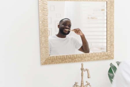 Happy African American man brushing his teeth with a toothbrush in front of a mirror in the morning...