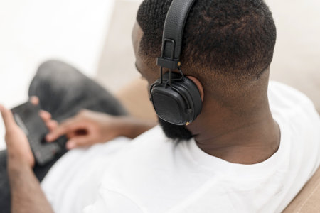African american young male enjoying music in wireless headphones while sitting on sofa at home. Close upの写真素材