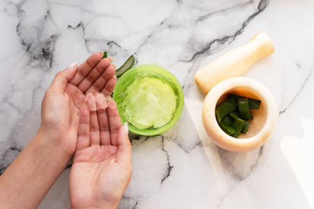 Hands of woman holding aloe gel next to mortar and chopped aloe leafの写真素材