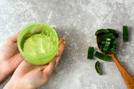 Female hands with aloe gel and a spoon with aloe leaf slices on a gray background. Making natural cosmetics at homeの写真素材