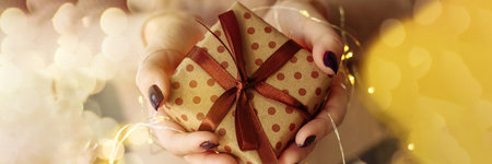 Hands hold and give a gift from craft brown packaging. A girl in a pink knitted sweater holds a New Year, Christmas present with a garland with yellow lights. Web bannerの写真素材