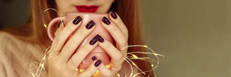 A young woman holds coffee near her mouth, drinks a warm drink in autumn or winter. Garland lights. Web banner, copy spaceの写真素材