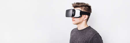 A young guy, a teenager stands in a villard vr glasses on a white background. Augmented reality in our time. A virtual reality glasses boy plays games and learns at school. Web banner.の写真素材