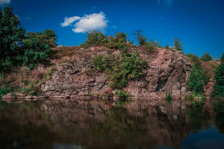 Landscape of red rocks by the river with green trees and blue sky.