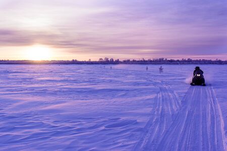 A snowmobile rides along the snowy ice of the river in the light of the setting sun.の写真素材