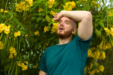 Portrait of handsome young allergic man is suffering from pollen allergy or cold on natural flowers,...