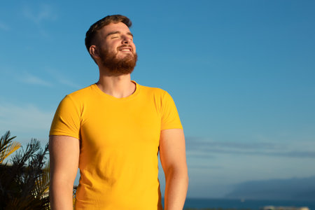 Portrait of happy cheerful positive guy, young handsome bearded man with beard is enjoying sunny day, summer vacation, sunbathing, tanning at sun and smiling, breathing deep, inhale fresh airの写真素材