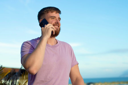 Happy young handsome man is talking on cell mobile phone, calling on smartphone, having conversation with friend, smiling outdoors at summer sunny dayの写真素材