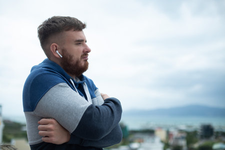 Portrait of bearded handsome pensive thoughtful guy, listen music in earphones, young upset sad frustrated man on natural mountain background, thinking with a serious faceの写真素材