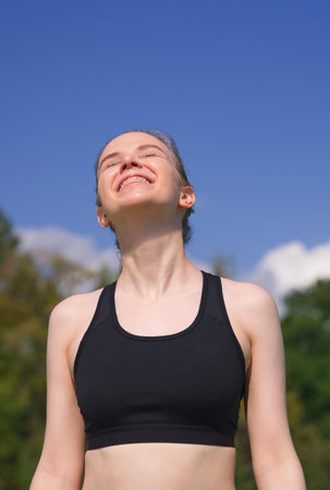 Happy positive fit fitness girl, joyful young woman is enjoying sun, good summer weather, sunbathing at sunny day on natural green and sky background. Vertical photo.の写真素材