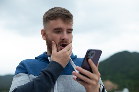 Angry or unhappy young man looking at the screen of the phone in the phone, the guy is surprised, upset, excitedの写真素材