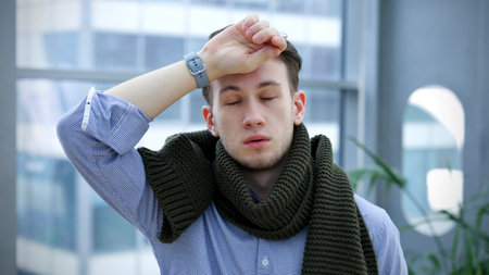 Young handsome exhausted ill sick man tired worker at office is suffering from heat, high temperature, headache and migraine, holding hand on forehead.の写真素材