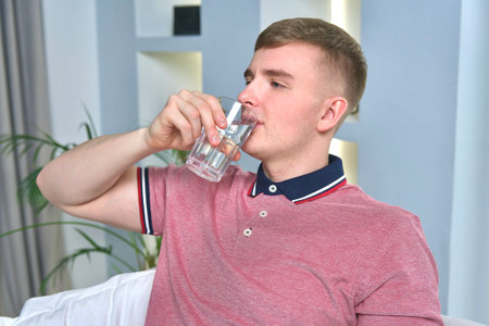 Young man is drinking pure fresh water from glass at home in living room